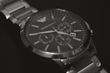 A silver luxury watch with a metallic strap and a dark face, featuring multiple dials for additional functions. The watch has a minimalist design with clear time markers and elegant branding visible on the dial.