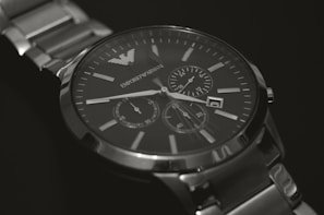 A silver luxury watch with a metallic strap and a dark face, featuring multiple dials for additional functions. The watch has a minimalist design with clear time markers and elegant branding visible on the dial.