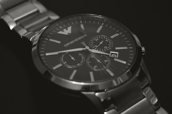 A silver luxury watch with a metallic strap and a dark face, featuring multiple dials for additional functions. The watch has a minimalist design with clear time markers and elegant branding visible on the dial.