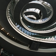 An architectural spiral staircase incorporating curves and glass elements, creating a modern and dynamic interior design. The structure spirals upwards with a combination of metal railings and glass panels, reflecting light and shadow playfully.