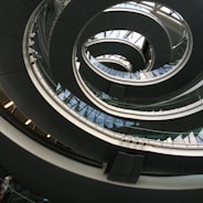 An architectural spiral staircase incorporating curves and glass elements, creating a modern and dynamic interior design. The structure spirals upwards with a combination of metal railings and glass panels, reflecting light and shadow playfully.
