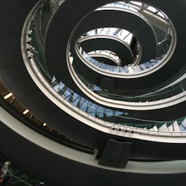 An architectural spiral staircase incorporating curves and glass elements, creating a modern and dynamic interior design. The structure spirals upwards with a combination of metal railings and glass panels, reflecting light and shadow playfully.