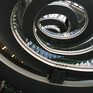 An architectural spiral staircase incorporating curves and glass elements, creating a modern and dynamic interior design. The structure spirals upwards with a combination of metal railings and glass panels, reflecting light and shadow playfully.