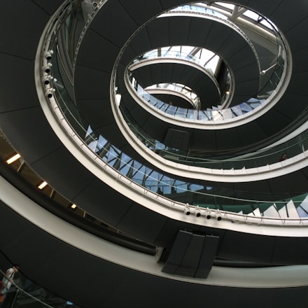An architectural spiral staircase incorporating curves and glass elements, creating a modern and dynamic interior design. The structure spirals upwards with a combination of metal railings and glass panels, reflecting light and shadow playfully.
