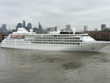 A large, white cruise ship named 'Silver Wind' is sailing along a river with a cityscape featuring modern skyscrapers and buildings in the background. The water reflects the cloudy sky, adding to the overcast atmosphere.