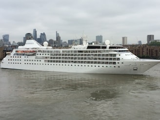 A large, white cruise ship named 'Silver Wind' is sailing along a river with a cityscape featuring modern skyscrapers and buildings in the background. The water reflects the cloudy sky, adding to the overcast atmosphere.