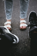 Two pairs of feet facing each other on a rough, gray asphalt surface. One person is wearing black athletic sneakers with white details, while the other is wearing white sandals over bare feet and gray jeans.
