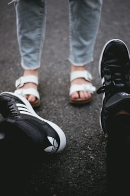 Two pairs of feet facing each other on a rough, gray asphalt surface. One person is wearing black athletic sneakers with white details, while the other is wearing white sandals over bare feet and gray jeans.