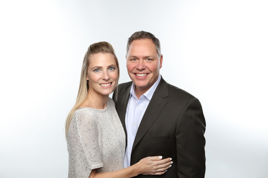 A smiling couple poses together, with the woman on the left wearing a light-colored textured sweater and the man on the right in a dark suit jacket and light blue shirt. The background is plain and white.