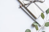 A neatly wrapped gift tied with a dark ribbon is placed next to a silver pencil. Green eucalyptus leaves lie beside them on a light, textured surface.