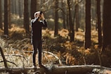 A person is standing on a fallen tree trunk in a dense forest, using a camera to take photographs. Sunlight filters through the trees, creating a warm and serene atmosphere with dappled light on the forest floor.