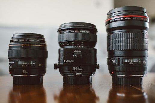 Three camera lenses are neatly arranged on a reflective wooden surface. Each lens is distinct in design and size, labeled with specifications such as focal lengths and apertures. The background is softly out of focus, highlighting the lenses in the foreground.