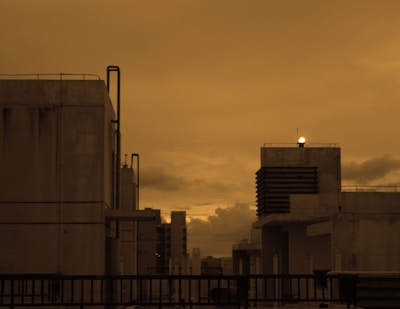 An industrial rooftop series of buildings silhouetted against a cloudy sky with a warm sepia tone. The structures feature piping and vents, contributing to an urban industrial atmosphere.