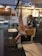 A butcher shop interior with a butcher working at a wooden table. A large piece of meat is hanging from a hook in the foreground. The shop has tiled walls, wooden cabinets, and various equipment such as scales, cutting boards, and a microwave. Bright overhead lights illuminate the space.