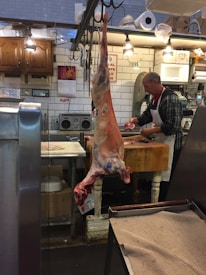 A butcher shop interior with a butcher working at a wooden table. A large piece of meat is hanging from a hook in the foreground. The shop has tiled walls, wooden cabinets, and various equipment such as scales, cutting boards, and a microwave. Bright overhead lights illuminate the space.