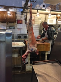 A butcher shop interior with a butcher working at a wooden table. A large piece of meat is hanging from a hook in the foreground. The shop has tiled walls, wooden cabinets, and various equipment such as scales, cutting boards, and a microwave. Bright overhead lights illuminate the space.