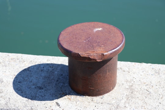 A rusted metal bollard is mounted on a concrete surface by a body of water. The bollard casts a distinct shadow, indicating direct sunlight. It appears weathered, with visible marks and discoloration.