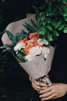 A bouquet of flowers wrapped in light brown paper, featuring orange and white blooms with green leaves. The bouquet is held by a pair of hands, with a leafy green plant in the background.