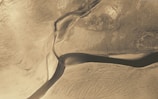 An aerial view of a meandering river splitting a vast landscape with intricate patterns formed by land and water erosion. The terrain appears arid with hues of brown and beige.