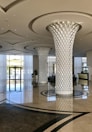 A modern and spacious hotel lobby featuring two large intricately designed columns with a leaf-like pattern. The area is bright and airy, with a high ceiling and shiny, reflective marble floors. Large floor-to-ceiling windows allow natural light to flood the space, and there is a reception desk in the background. Potted plants add a touch of greenery.