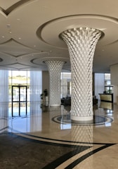 A modern and spacious hotel lobby featuring two large intricately designed columns with a leaf-like pattern. The area is bright and airy, with a high ceiling and shiny, reflective marble floors. Large floor-to-ceiling windows allow natural light to flood the space, and there is a reception desk in the background. Potted plants add a touch of greenery.
