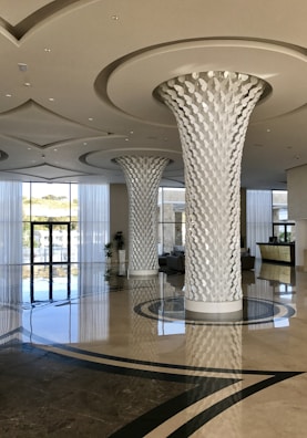 A modern and spacious hotel lobby featuring two large intricately designed columns with a leaf-like pattern. The area is bright and airy, with a high ceiling and shiny, reflective marble floors. Large floor-to-ceiling windows allow natural light to flood the space, and there is a reception desk in the background. Potted plants add a touch of greenery.