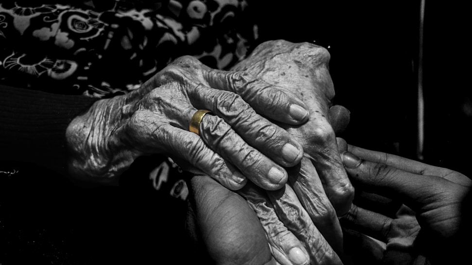 Two hands are clasped together, with one appearing more aged and the other younger. A golden ring is worn on one finger of the older hand. The background is dark, highlighting the contrast between the wrinkles on the older hand and the smoother skin of the younger one.