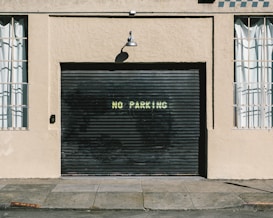A black garage door with the words 'NO PARKING' painted in white is set into a beige stucco building. On either side of the door are windows with white curtains and metal bars. Above the garage is an industrial-style metal lamp casting a shadow on the wall.