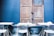 A rustic wooden door is flanked by a dark blue wall, creating a striking contrast. In front of the door, modern white metal chairs and tables are arranged neatly. Cushions are placed on a narrow bench against the wall, adding a touch of comfort to the outdoor seating area.