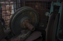A close-up view of rusty, old mechanical parts, including wheels and axles, surrounded by a metal grid or cage. The surface of the metal components displays signs of wear and corrosion, with a dark and gritty texture.