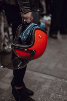 A person wearing a black leather jacket holds a red motorcycle helmet in their hand. A blue cloth is tucked inside the helmet, and the person is wearing dark pants and black boots. The focus is on the helmet and hand, while the background is blurred.