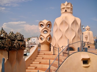 An architectural rooftop scene featuring uniquely shaped chimneys and ventilation towers with organic, flowing designs reminiscent of a wave pattern. The structures are made of a light, earthy colored stone and contrast with the clear blue sky. The foreground includes a winding staircase with a metal railing, leading to different levels of the rooftop.