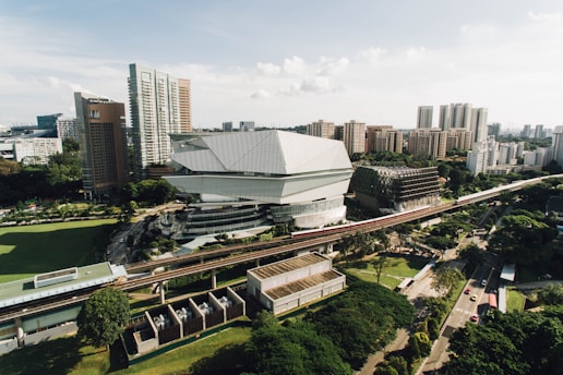 A modern urban landscape featuring a distinctive architectural building with sharp, angular designs surrounded by high-rise buildings. The area includes a monorail track running through the scene, with lush greenery scattered throughout the surroundings.