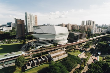 A modern urban landscape featuring a distinctive architectural building with sharp, angular designs surrounded by high-rise buildings. The area includes a monorail track running through the scene, with lush greenery scattered throughout the surroundings.