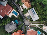 An aerial view of a suburban neighborhood featuring several houses with red and gray roofs. The properties are surrounded by lush greenery and well-maintained gardens. Two swimming pools are visible in the backyards of two different houses. A road runs through the center of the image, separating the houses and bordered by trees.