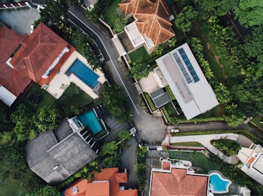 An aerial view of a suburban neighborhood featuring several houses with red and gray roofs. The properties are surrounded by lush greenery and well-maintained gardens. Two swimming pools are visible in the backyards of two different houses. A road runs through the center of the image, separating the houses and bordered by trees.