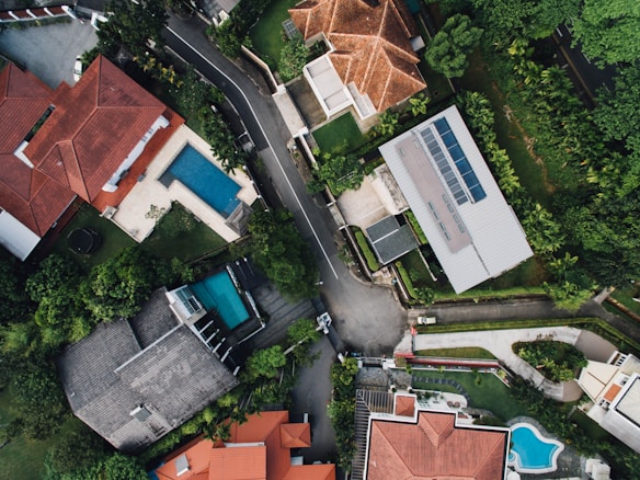 An aerial view of a suburban neighborhood featuring several houses with red and gray roofs. The properties are surrounded by lush greenery and well-maintained gardens. Two swimming pools are visible in the backyards of two different houses. A road runs through the center of the image, separating the houses and bordered by trees.