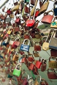 Numerous colorful padlocks attached to a wire mesh fence, with some padlocks inscribed with names and messages. The focus is on the locks, suggesting themes of love and commitment.