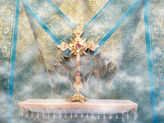 A decorative and ornate golden cross studded with red jewels is placed on an intricately designed blue and gold patterned fabric background. The cross stands on a small, white altar with fringe detail, enveloped in a soft mist that adds an ethereal quality to the scene.
