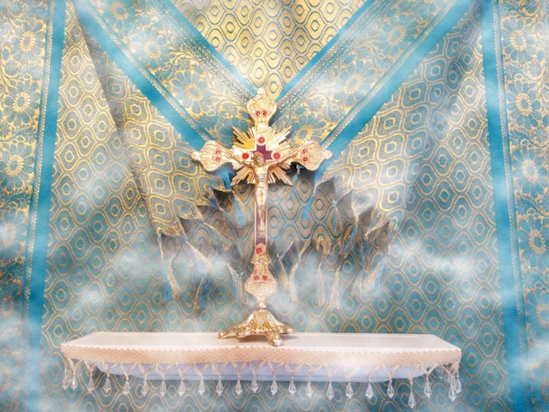 A decorative and ornate golden cross studded with red jewels is placed on an intricately designed blue and gold patterned fabric background. The cross stands on a small, white altar with fringe detail, enveloped in a soft mist that adds an ethereal quality to the scene.
