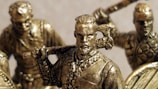 Bronze figurines of warriors are depicted, featuring detailed armor and weapons like axes and swords. The figures appear to be in dynamic poses, suggesting action or readiness for battle.