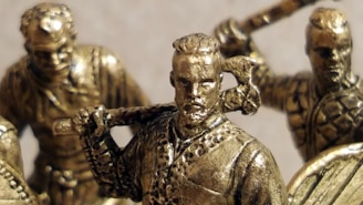 Bronze figurines of warriors are depicted, featuring detailed armor and weapons like axes and swords. The figures appear to be in dynamic poses, suggesting action or readiness for battle.
