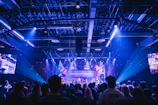 A wide shot of a concert stage illuminated with deep blue and gold lights, filled with a lively crowd.