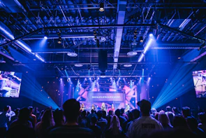 A wide shot of a concert stage illuminated with deep blue and gold lights, filled with a lively crowd.