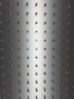 Metallic surface with evenly spaced small circular holes in a grid pattern. Light gradient transitioning from darker tones at the edges to a brighter center, giving a three-dimensional effect.