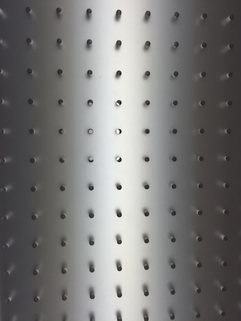Metallic surface with evenly spaced small circular holes in a grid pattern. Light gradient transitioning from darker tones at the edges to a brighter center, giving a three-dimensional effect.