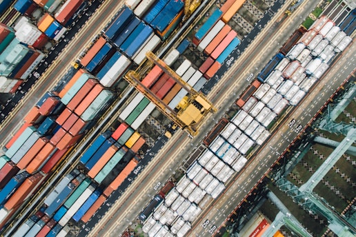 An aerial view of a busy shipping port filled with numerous colorful shipping containers. The scene includes various sizes and colors of containers arranged in neat rows, with cranes and vehicles for transportation. The layout is orderly and structured, indicating a highly organized operation.