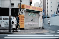 An urban street scene featuring a closed shop with a corrugated metal shutter displaying 'BEST SERVICE' in faded lettering. The shop is surrounded by buildings, and there are red, yellow, and white signs with Japanese text. Two individuals are walking on the sidewalk next to the shop, and another person is walking away on the crosswalk. The street is quiet with minimal traffic.