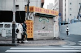An urban street scene featuring a closed shop with a corrugated metal shutter displaying 'BEST SERVICE' in faded lettering. The shop is surrounded by buildings, and there are red, yellow, and white signs with Japanese text. Two individuals are walking on the sidewalk next to the shop, and another person is walking away on the crosswalk. The street is quiet with minimal traffic.