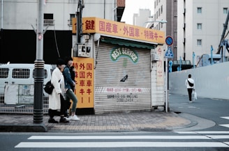 An urban street scene featuring a closed shop with a corrugated metal shutter displaying 'BEST SERVICE' in faded lettering. The shop is surrounded by buildings, and there are red, yellow, and white signs with Japanese text. Two individuals are walking on the sidewalk next to the shop, and another person is walking away on the crosswalk. The street is quiet with minimal traffic.